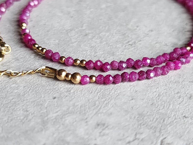 Pu&ograve; includere: Primo piano di una delicata collana con piccole pietre preziose rosa intenso sfaccettate e perline color oro. La collana ha una chiusura e una catena color oro. Il gioiello &egrave; posto su uno sfondo strutturato bianco sporco.