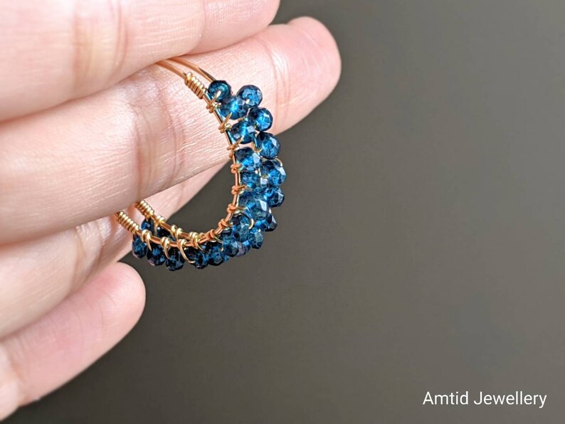 May include: Gold wire hoop earrings with blue gemstone beads. The earrings are handmade and feature a cluster of small, round blue gemstones.