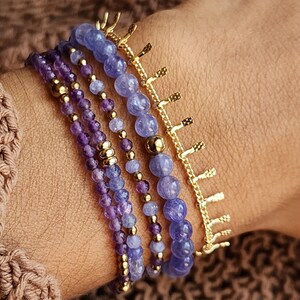 May include: A set of five bracelets featuring amethyst and blue gemstone beads with gold accents. The bracelets are layered on a person's wrist.