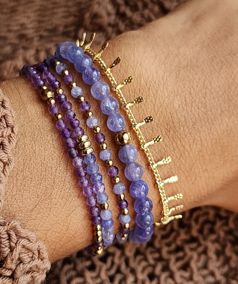 May include: A set of five bracelets featuring amethyst and blue gemstone beads with gold accents. The bracelets are layered on a person's wrist.
