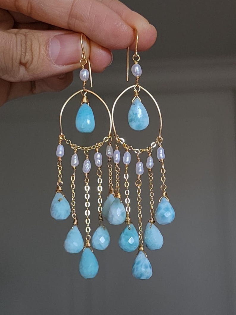 May include: Gold chandelier earrings with light blue teardrop gemstones and small white pearls. The earrings have a delicate chain design, suspended from a gold hook. The gemstones are approximately 1.5 cm in length.