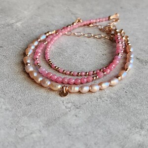 Puede incluir: Tres pulseras en capas con detalles en rosa y dorado. La pulsera superior presenta peque&ntilde;as cuentas rosas, la del medio tiene cuentas rosas m&aacute;s grandes y la inferior perlas ovaladas. Un peque&ntilde;o dije dorado cuelga de la pulsera inferior.