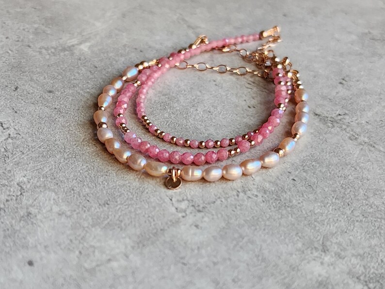 Puede incluir: Tres pulseras en capas con detalles en rosa y dorado. La pulsera superior presenta peque&ntilde;as cuentas rosas, la del medio tiene cuentas rosas m&aacute;s grandes y la inferior perlas ovaladas. Un peque&ntilde;o dije dorado cuelga de la pulsera inferior.