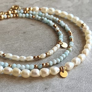 May include: Three layered bracelets featuring freshwater pearls, light blue beads, and gold-colored accents. The bracelets have a delicate design with small gold beads and round gold charms. The pearls are off-white and the beads are a light blue color.