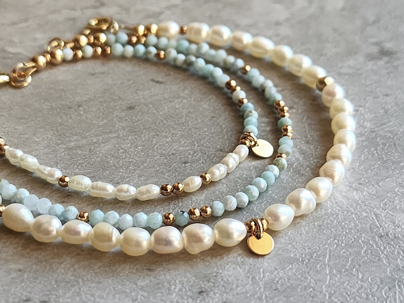 May include: Three layered bracelets featuring freshwater pearls, light blue beads, and gold-colored accents. The bracelets have a delicate design with small gold beads and round gold charms. The pearls are off-white and the beads are a light blue color.