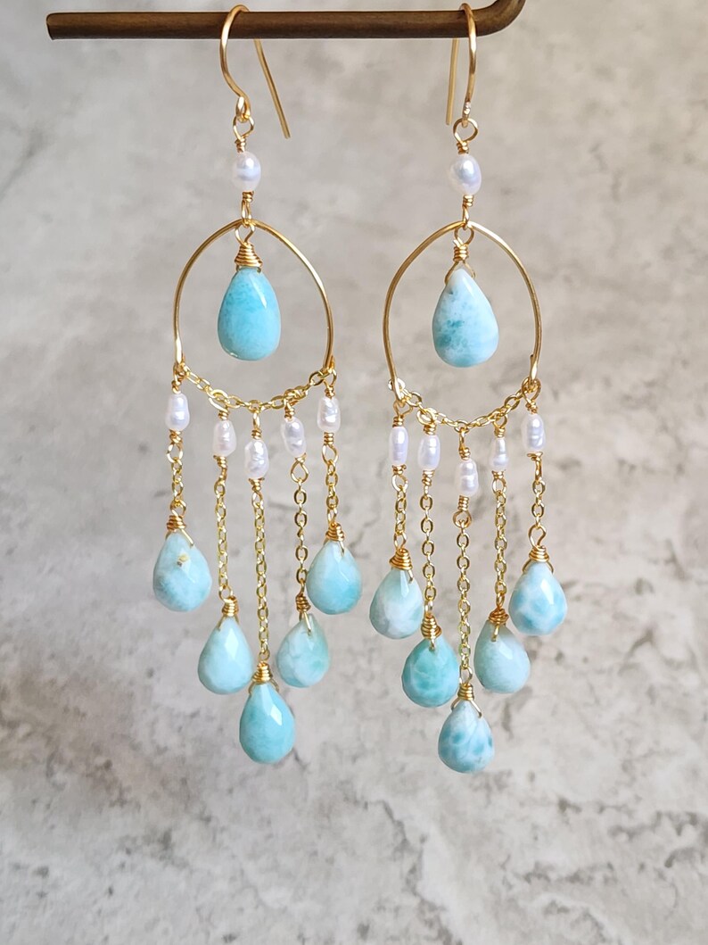 May include: Gold-tone chandelier earrings with teardrop-shaped, light blue gemstones and small white pearls. The earrings have a delicate chain design and are suspended from gold-tone hooks.