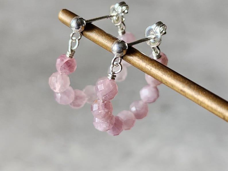 May include: A pair of silver hoop earrings with pink gemstone beads. The earrings are hanging from a wooden display stand.