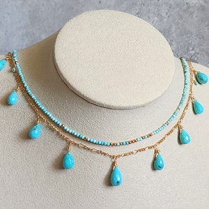 May include: A two-strand necklace displayed on a mannequin. The top strand is made of small turquoise beads, while the bottom strand is a gold chain. Turquoise teardrop-shaped pendants hang from the chains, creating a layered effect.