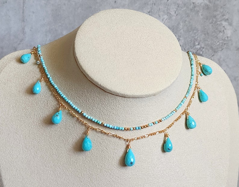 May include: A two-strand necklace displayed on a mannequin. The top strand is made of small turquoise beads, while the bottom strand is a gold chain. Turquoise teardrop-shaped pendants hang from the chains, creating a layered effect.
