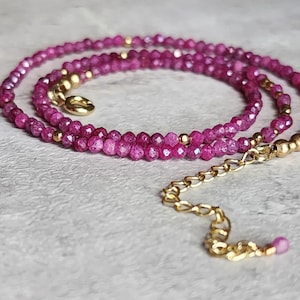 May include: A delicate necklace with small, faceted, magenta beads. It has gold-coloured accents and a gold-coloured chain with a lobster clasp. The necklace is designed to be worn around the neck.