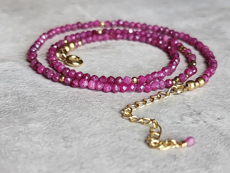 Pu&ograve; includere: Una delicata collana con piccole perline sfaccettate color magenta. La collana &egrave; impreziosita da perline color oro e una chiusura a catena color oro. La collana &egrave; esposta su una superficie grigio chiaro.
