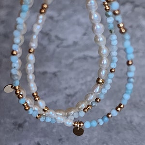 May include: Three layered bracelets featuring freshwater pearls, light blue beads, and gold-colored accents. Each bracelet has a different design, with a mix of bead sizes and textures. Small gold-colored disc charms dangle from the bracelets.