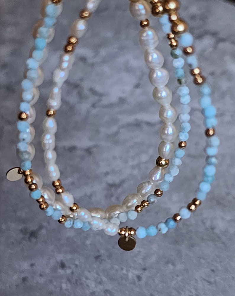 May include: Three layered bracelets featuring freshwater pearls, light blue beads, and gold-colored accents. Each bracelet has a different design, with a mix of bead sizes and textures. Small gold-colored disc charms dangle from the bracelets.