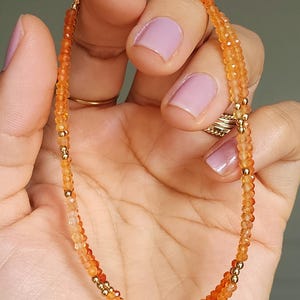 May include: A delicate orange gemstone bracelet with gold-colored beads. The bracelet is composed of small, faceted orange beads strung together, interspersed with small gold-colored beads. The bracelet is held in a hand with painted nails.