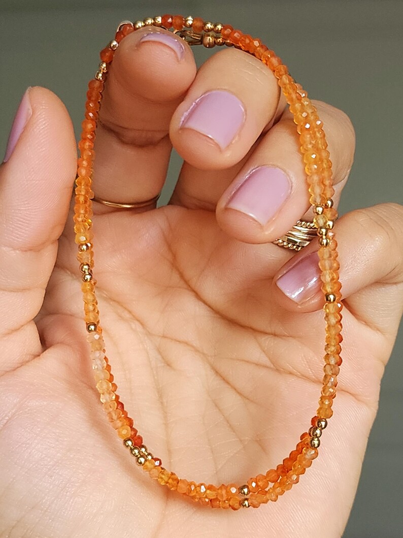 May include: A delicate orange gemstone bracelet with gold-colored beads. The bracelet is composed of small, faceted orange beads strung together, interspersed with small gold-colored beads. The bracelet is held in a hand with painted nails.