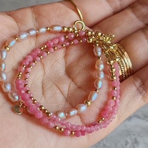 Puede incluir: Pulsera delicada con tres hilos. Un hilo tiene piedras preciosas rosas, otro perlas blancas y el tercero piedras preciosas rosas. Cuentas y detalles dorados se intercalan en la pulsera, que se sostiene en una mano.