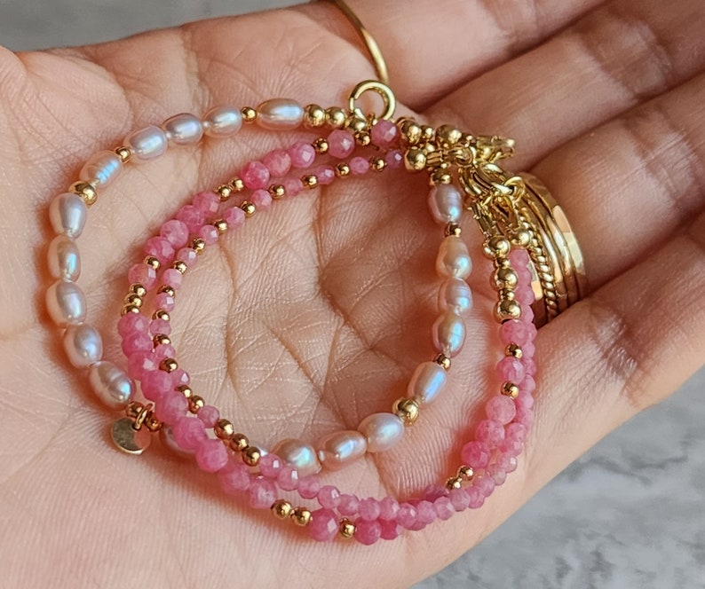 Puede incluir: Pulsera delicada con tres hilos. Un hilo tiene piedras preciosas rosas, otro perlas blancas y el tercero piedras preciosas rosas. Cuentas y detalles dorados se intercalan en la pulsera, que se sostiene en una mano.