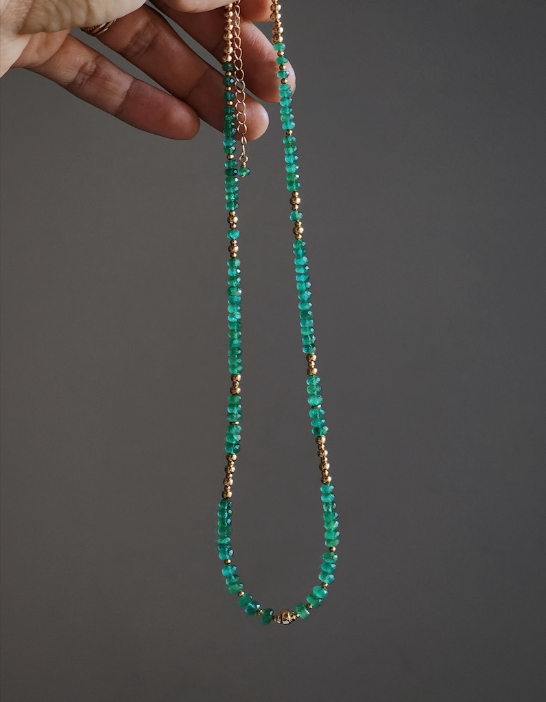 May include: A delicate gold chain necklace with green gemstone beads. The necklace is made of small, round beads that are strung together to create a simple and elegant design.