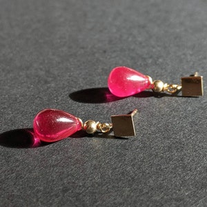 May include: A pair of dangle earrings with pink teardrop-shaped stones and gold square accents.