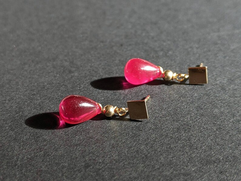 May include: A pair of dangle earrings with pink teardrop-shaped stones and gold square accents.