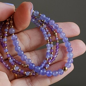 May include: A multi-strand bracelet featuring a gradient of purple and blue gemstone beads. The bracelet is made with small, round beads and has a gold accent.