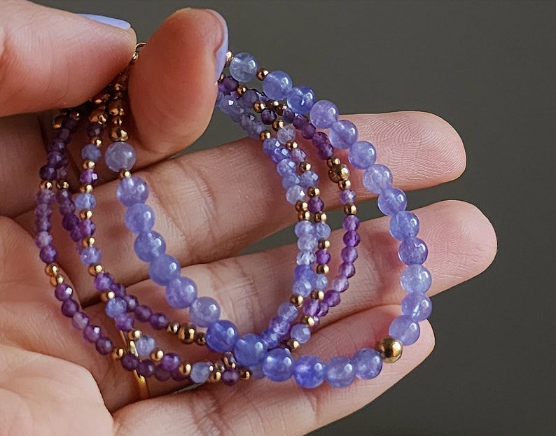 May include: A multi-strand bracelet featuring a gradient of purple and blue gemstone beads. The bracelet is made with small, round beads and has a gold accent.