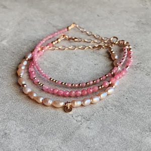Puede incluir: Pulsera de tres hilos con piedras preciosas rosas, perlas y cadena dorada. La pulsera tiene un dise&ntilde;o delicado con un peque&ntilde;o dije redondo. Las perlas son de color blanco cremoso y las piedras preciosas son de un rosa vibrante.