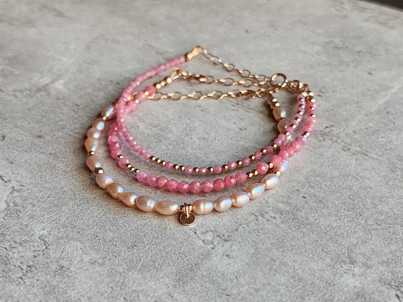 Puede incluir: Pulsera de tres hilos con piedras preciosas rosas, perlas y cadena dorada. La pulsera tiene un dise&ntilde;o delicado con un peque&ntilde;o dije redondo. Las perlas son de color blanco cremoso y las piedras preciosas son de un rosa vibrante.