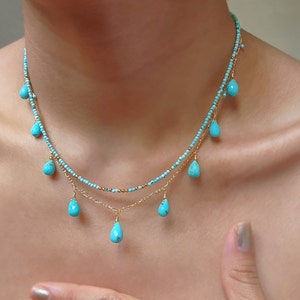 May include: A delicate, layered necklace featuring small turquoise beads and teardrop-shaped turquoise pendants. The necklace has gold-colored chain accents and is displayed on a person's neck. The jewelry is a light blue color.
