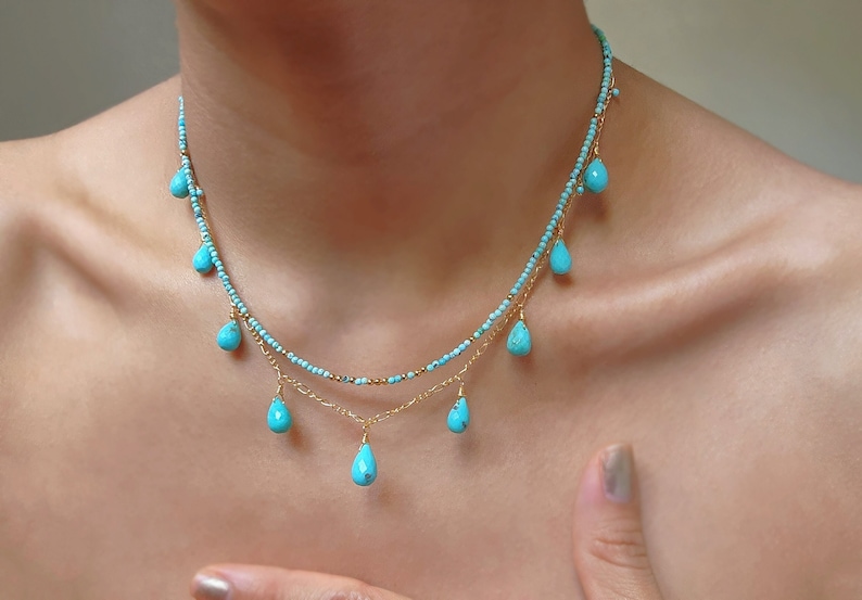 May include: A delicate, layered necklace featuring small turquoise beads and teardrop-shaped turquoise pendants. The necklace has gold-colored chain accents and is displayed on a person's neck. The jewelry is a light blue color.
