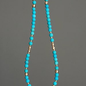 May include: A turquoise and gold beaded necklace with a gold bead accent. The necklace is made of small, round beads.