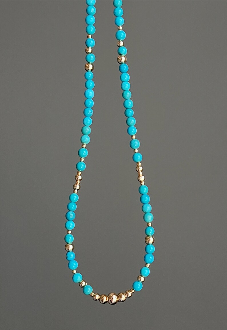 May include: A turquoise and gold beaded necklace with a gold bead accent. The necklace is made of small, round beads.