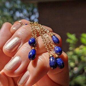 May include: Gold-toned chain earrings with multiple strands, each adorned with faceted, deep blue lapis lazuli teardrop beads. The earrings are held in a hand, showcasing the intricate design and color contrast.