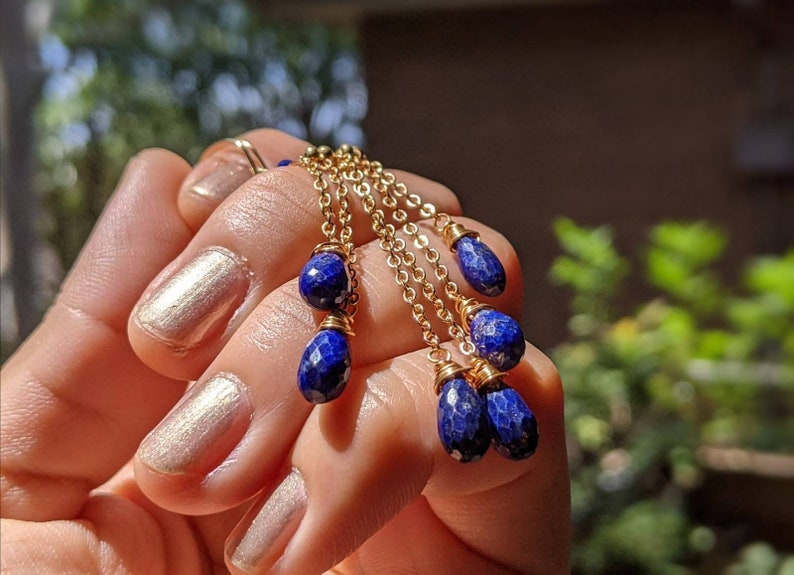 May include: Gold-toned chain earrings with multiple strands, each adorned with faceted, deep blue lapis lazuli teardrop beads. The earrings are held in a hand, showcasing the intricate design and color contrast.