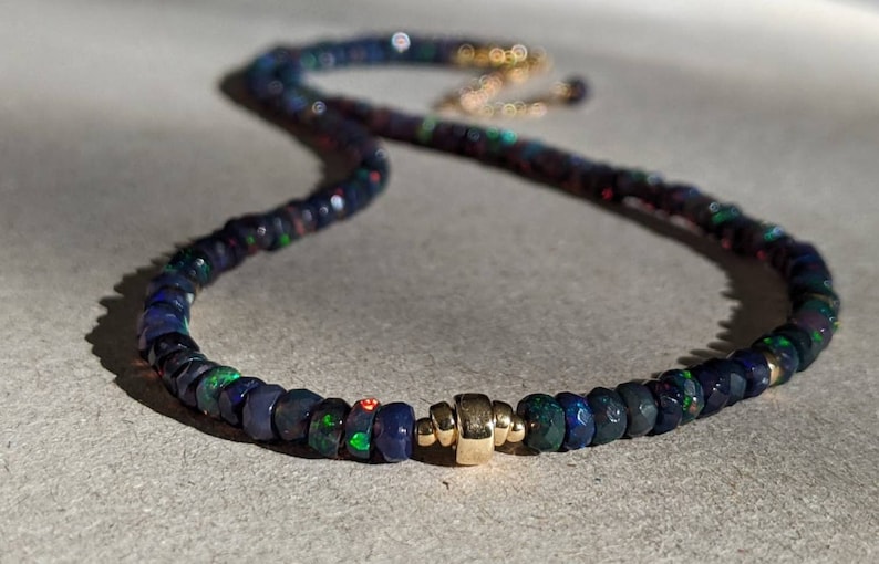 Op de afbeelding: Een ketting met gefacetteerde donkerblauwe en zwarte opaal kralen met iriserende groene en rode flitsen. De streng wordt geaccentueerd door een centrale gepolijste goudkleurige cilindrische kraal en heeft een goudkleurige metalen sluiting.