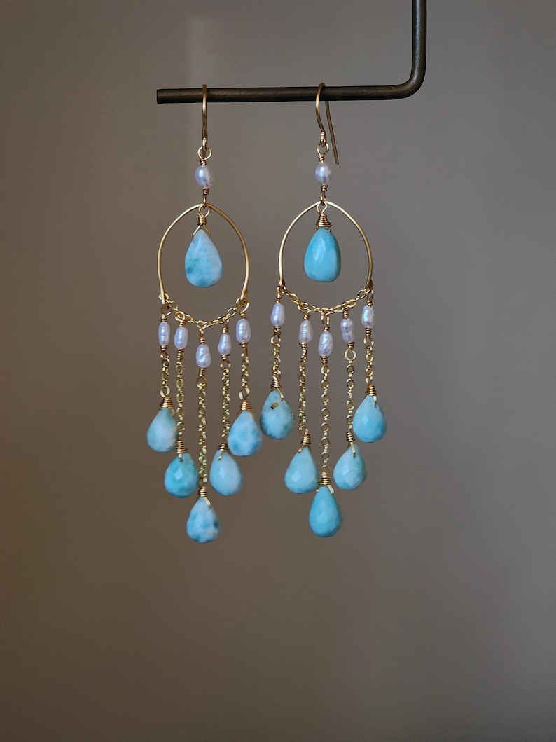 May include: A pair of gold-colored earrings with a teardrop-shaped, light blue stone suspended from a gold hoop. Small white pearls and additional light blue teardrop stones dangle from the hoop on gold chains. The earrings are suspended from a dark metal bar.