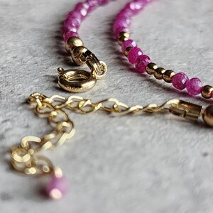 Pu&ograve; includere: Primo piano di una collana con piccole pietre preziose rosa sfaccettate e perline color oro. La collana ha una chiusura color oro e una catena di estensione. Il gioiello &egrave; esposto su una superficie strutturata grigio chiaro.