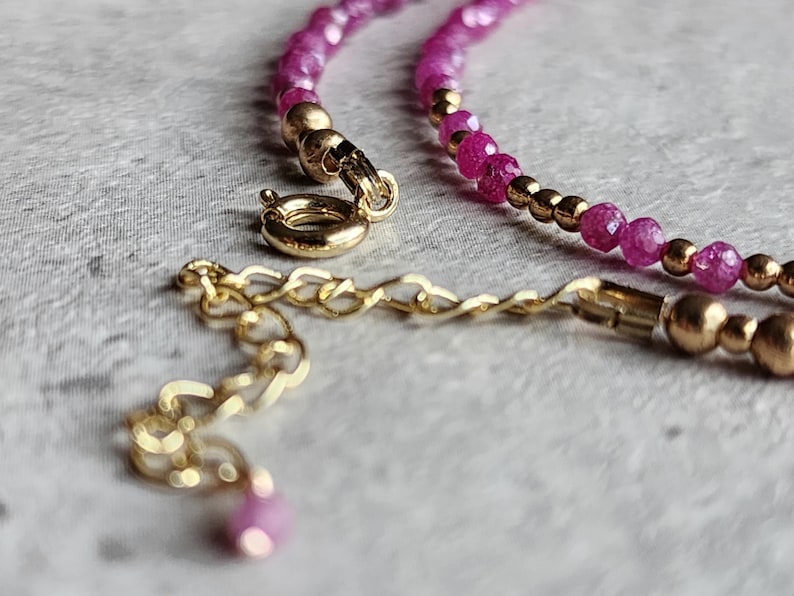 Pu&ograve; includere: Primo piano di una collana con piccole pietre preziose rosa sfaccettate e perline color oro. La collana ha una chiusura color oro e una catena di estensione. Il gioiello &egrave; esposto su una superficie strutturata grigio chiaro.