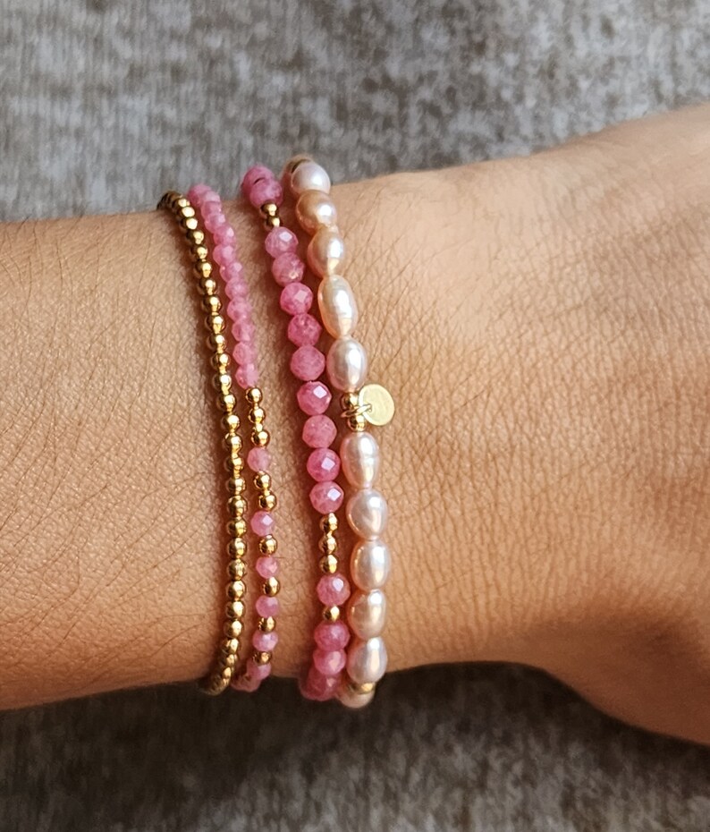 Puede incluir: Un conjunto de tres pulseras en tonos rosa y dorado. Una pulsera presenta peque&ntilde;as cuentas doradas, otra tiene cuentas de piedras preciosas rosas, y la tercera combina piedras rosas con perlas blancas. Se adjunta un peque&ntilde;o dije dorado.