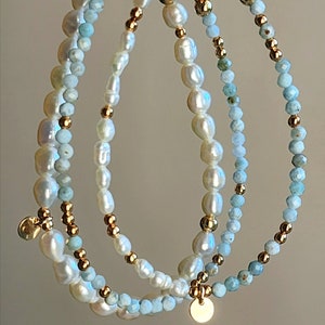 May include: Three delicate bracelets featuring freshwater pearls, light blue gemstones, and gold-colored beads. Each bracelet has a different design, with a small gold charm hanging from two of them. The bracelets are strung together.