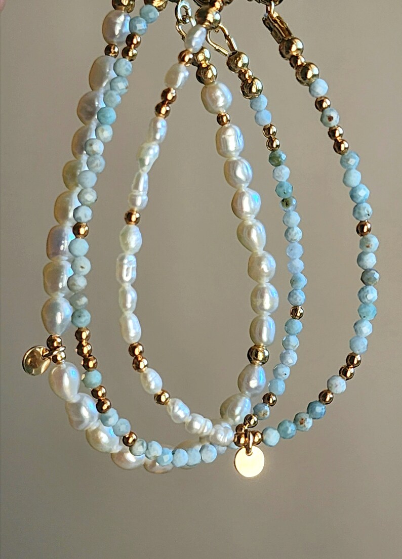 May include: Three delicate bracelets featuring freshwater pearls, light blue gemstones, and gold-colored beads. Each bracelet has a different design, with a small gold charm hanging from two of them. The bracelets are strung together.