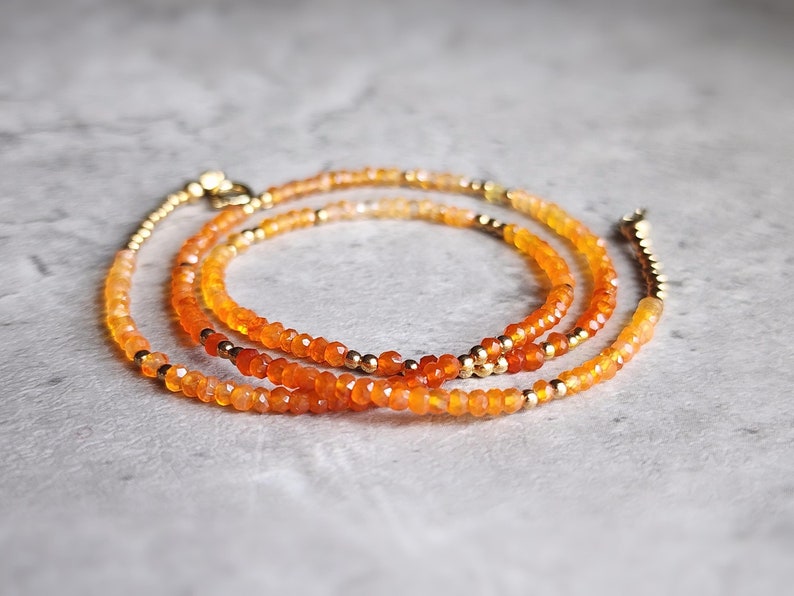 May include: A layered necklace featuring strands of orange and gold beads. The necklace has a delicate design with small, faceted orange beads and interspersed gold-colored beads. The clasp and connecting elements are also gold-toned.