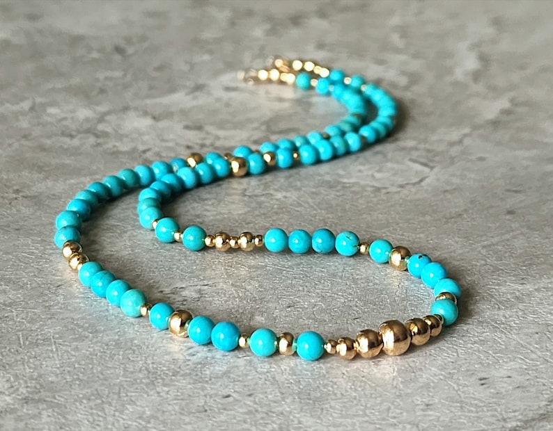 May include: A turquoise and gold beaded necklace. The necklace is made of small, round turquoise beads and gold beads. The necklace is on a gray surface.