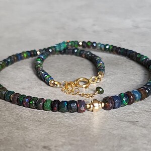 Op de afbeelding: Een ketting met kleine, gefacetteerde, donkergekleurde kralen met iriserende groene en blauwe flitsen. De streng is voorzien van een goudkleurige metalen sluiting en een korte verlengketting, wat zorgt voor een cirkelvormige opstelling.