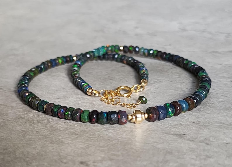 Op de afbeelding: Een ketting met kleine, gefacetteerde, donkergekleurde kralen met iriserende groene en blauwe flitsen. De streng is voorzien van een goudkleurige metalen sluiting en een korte verlengketting, wat zorgt voor een cirkelvormige opstelling.