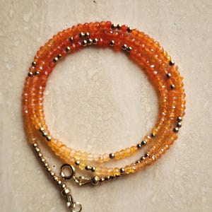 May include: A layered necklace featuring small, faceted orange and yellow beads. Gold-colored beads are interspersed throughout the necklace. The necklace has a gold-colored clasp.