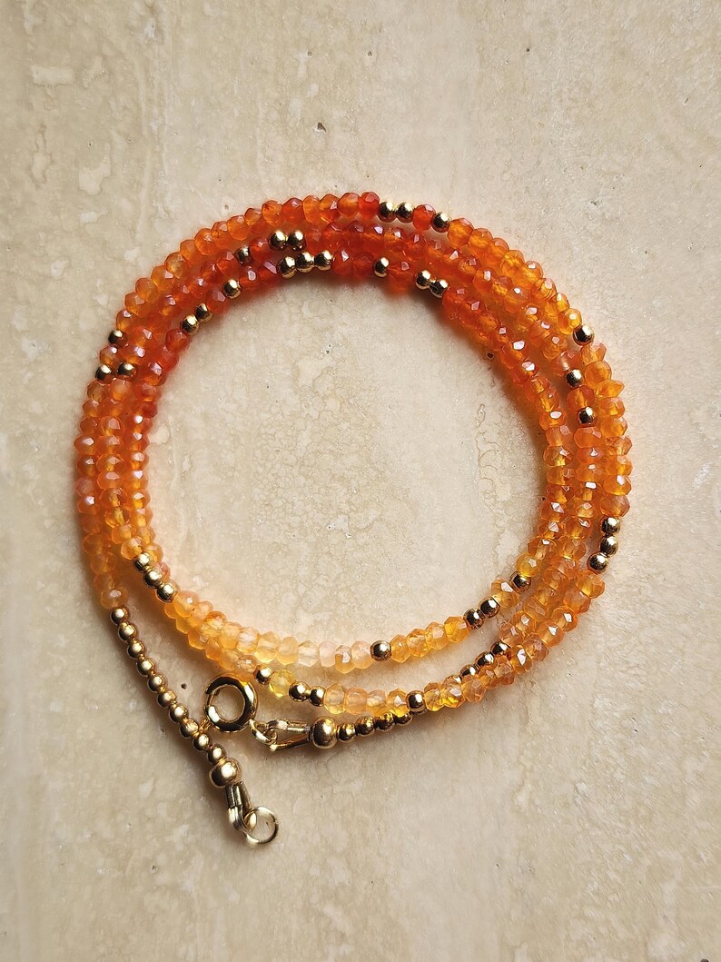 May include: A layered necklace featuring small, faceted orange and yellow beads. Gold-colored beads are interspersed throughout the necklace. The necklace has a gold-colored clasp.