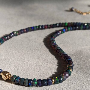 Op de afbeelding: Een kralenketting met kleine, gefacetteerde donkerblauwe en zwarte opaal kralen met iriserende groene en rode flitsen. De streng is geaccentueerd met gepolijste goudkleurige metalen tussenkralen en heeft een goudkleurige metalen sluiting.