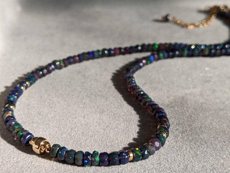 Op de afbeelding: Een kralenketting met kleine, gefacetteerde donkerblauwe en zwarte opaal kralen met iriserende groene en rode flitsen. De streng is geaccentueerd met gepolijste goudkleurige metalen tussenkralen en heeft een goudkleurige metalen sluiting.