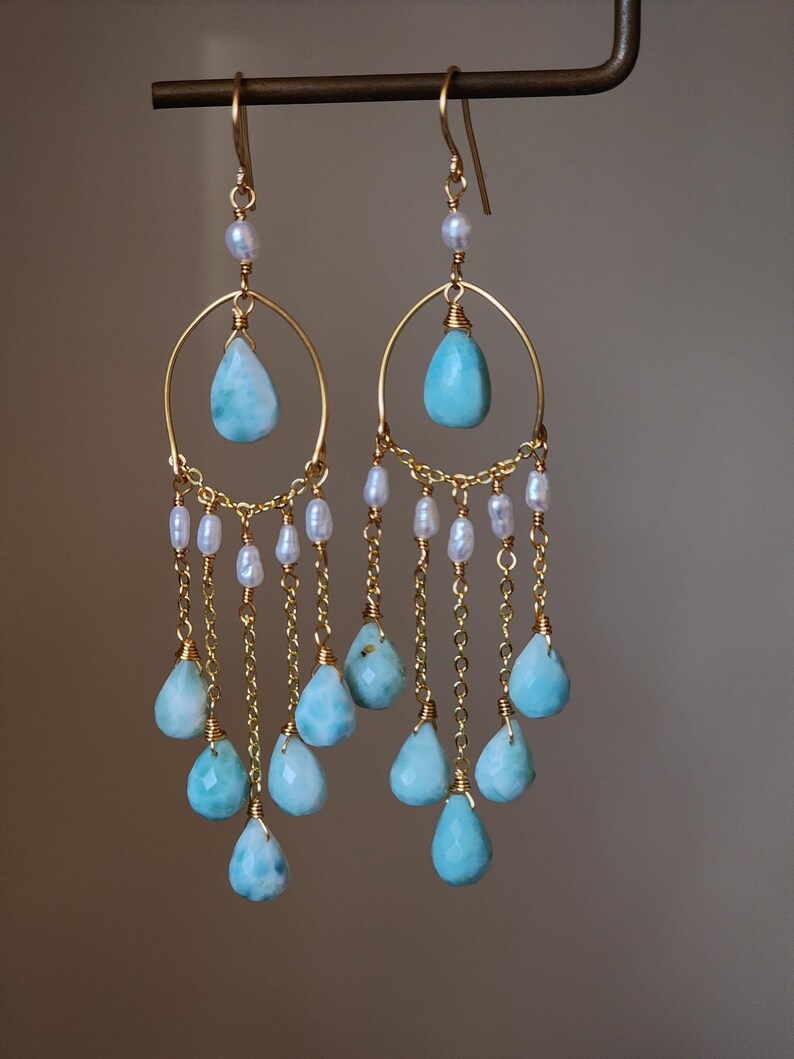 May include: A pair of gold-toned earrings featuring teardrop-shaped, light blue gemstones and small white pearls. The earrings have a chandelier design with multiple dangling gemstones suspended from delicate chains.